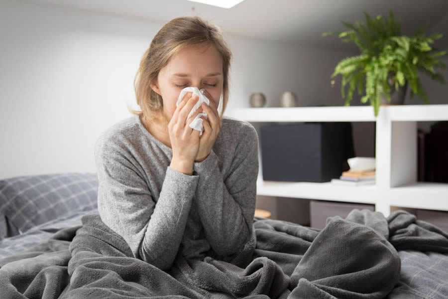 Mulher sentada na cama, com uma coberta nas pernas, assoando o nariz