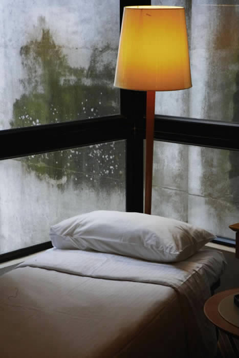 Quarto com uma cama e um abajur, envolto de vidro, e ao fundo uma parede com mofo